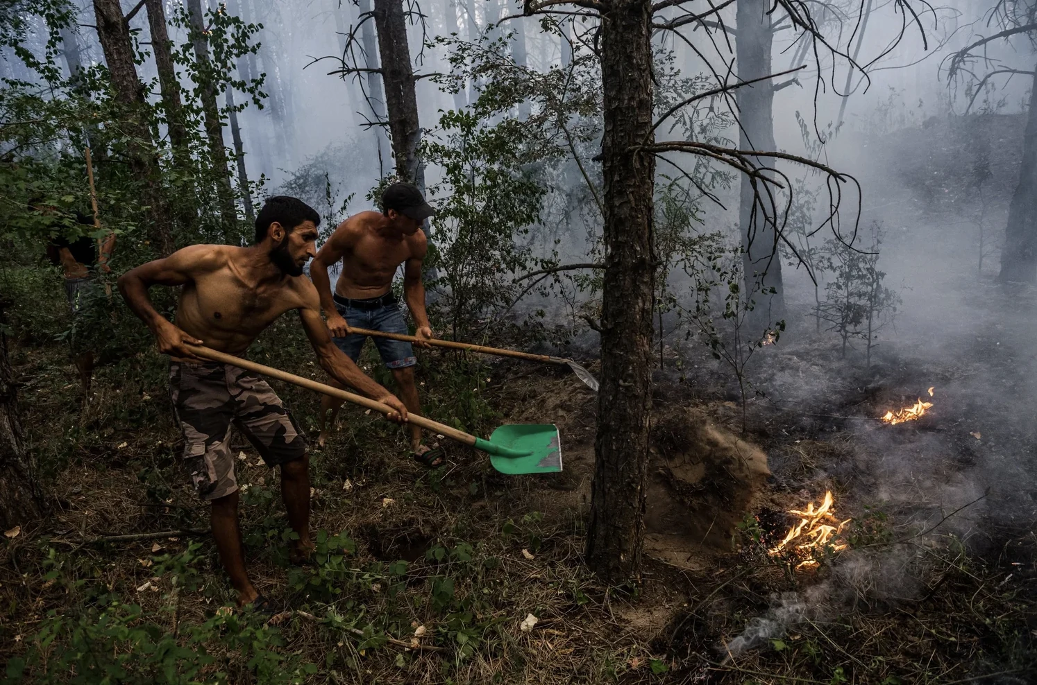 Two people using shovels near a fire in a smoke-filled forest